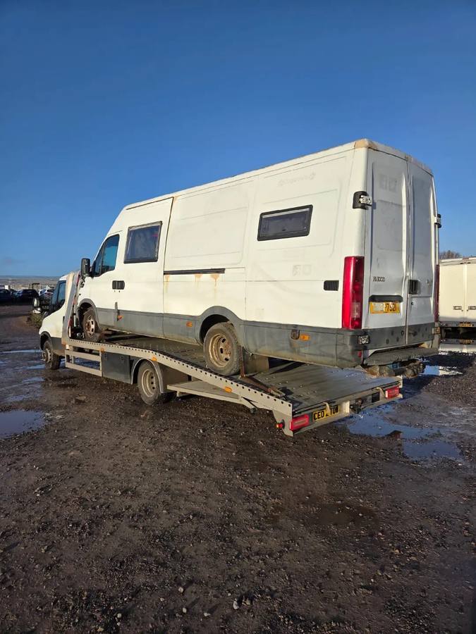 White Iveco van transported on recovery flatbed at muddy outdoor lot